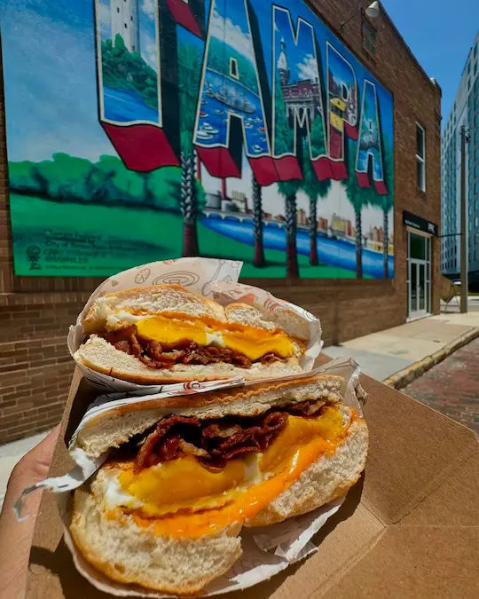 Three breakfast sandwiches from Tampa Bay cafés on a rustic table, showing different bread, fillings, and styles.